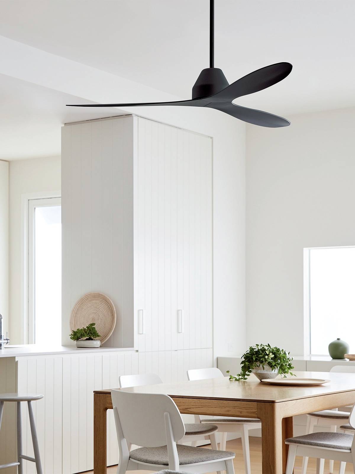 Een moderne, zwarte plafondventilator draait boven een eettafel met witte stoelen in een lichte, minimalistische ruimte.