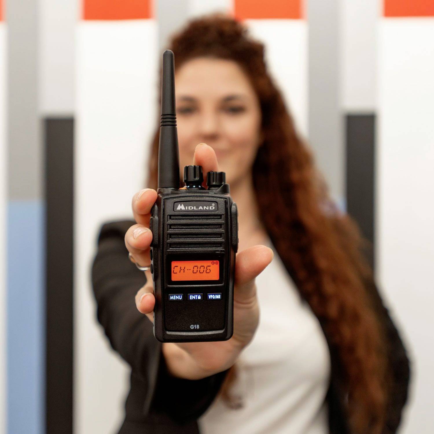 Een vrouw houdt een portofoon voor de camera. Het display toont 'CH-006'. Ze heeft lang, krullend haar en draagt een blazer.