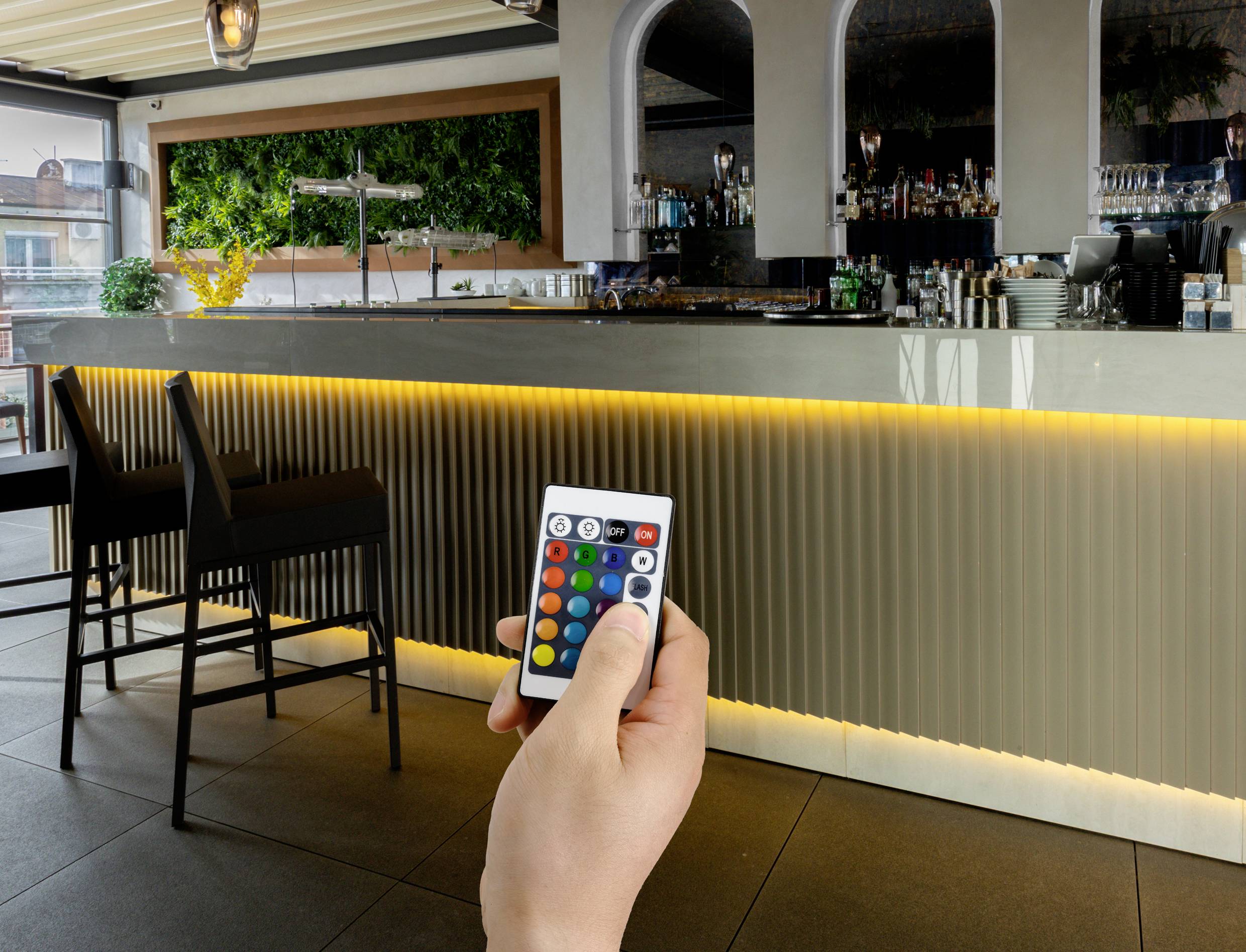 Een persoon richt een afstandsbediening op een moderne bar met verlichte toonbank en stoelen in het interieur.