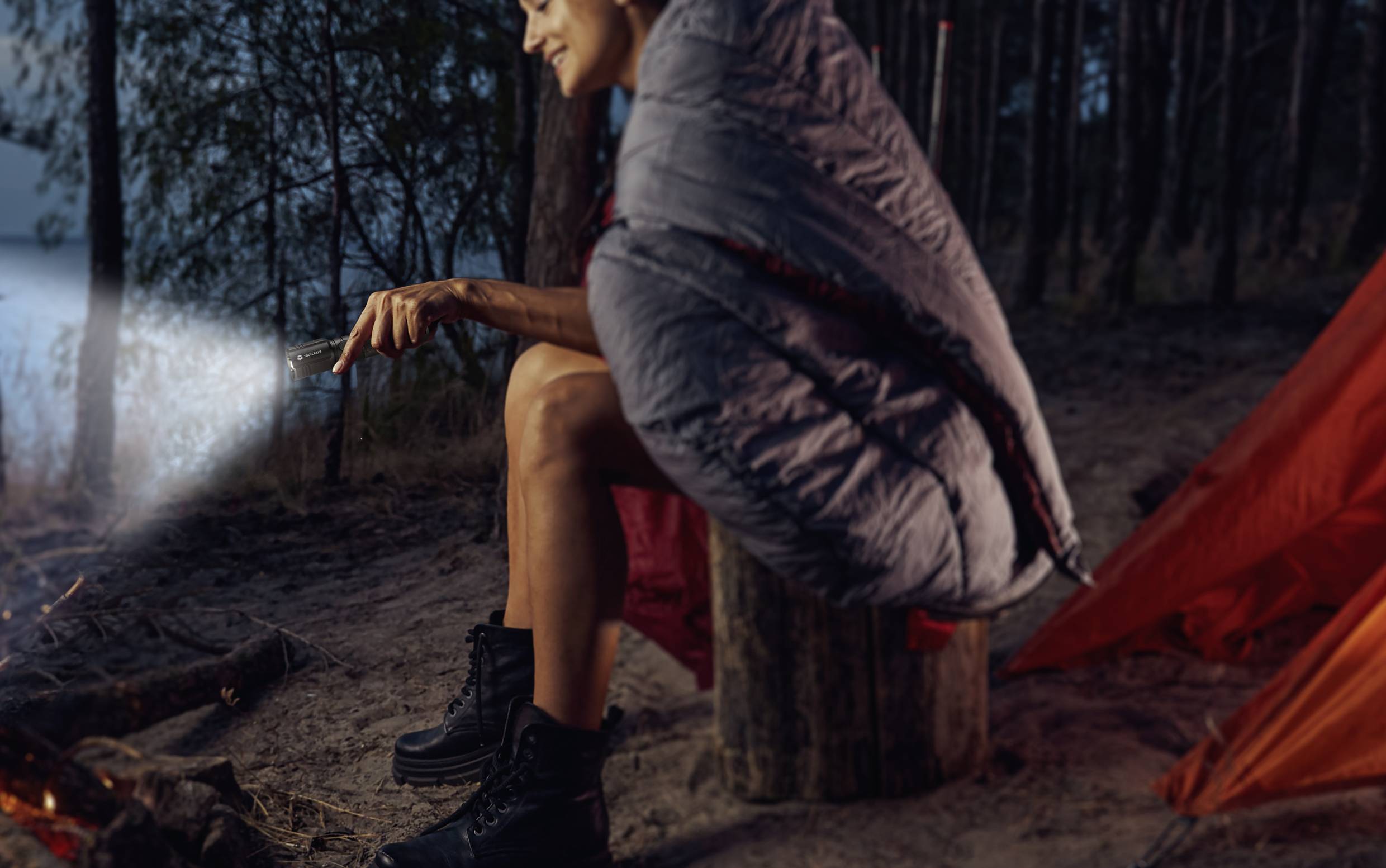 Een persoon zit in een slaapzak naast een kampvuur in het bos, houdt een zaklamp vast en glimlacht. Tent en bomen op de achtergrond.