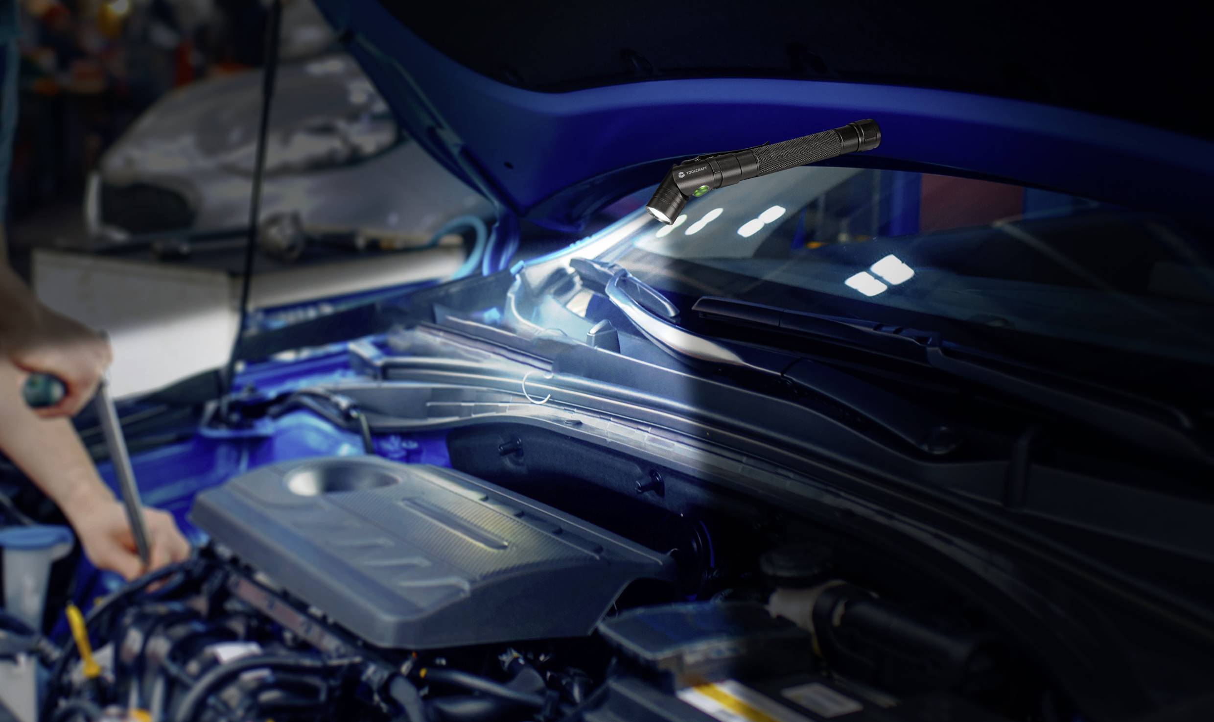 Een monteur werkt onder de opengeklapte motorkap van een auto, verlicht door een lamp. De focus ligt op technische details.