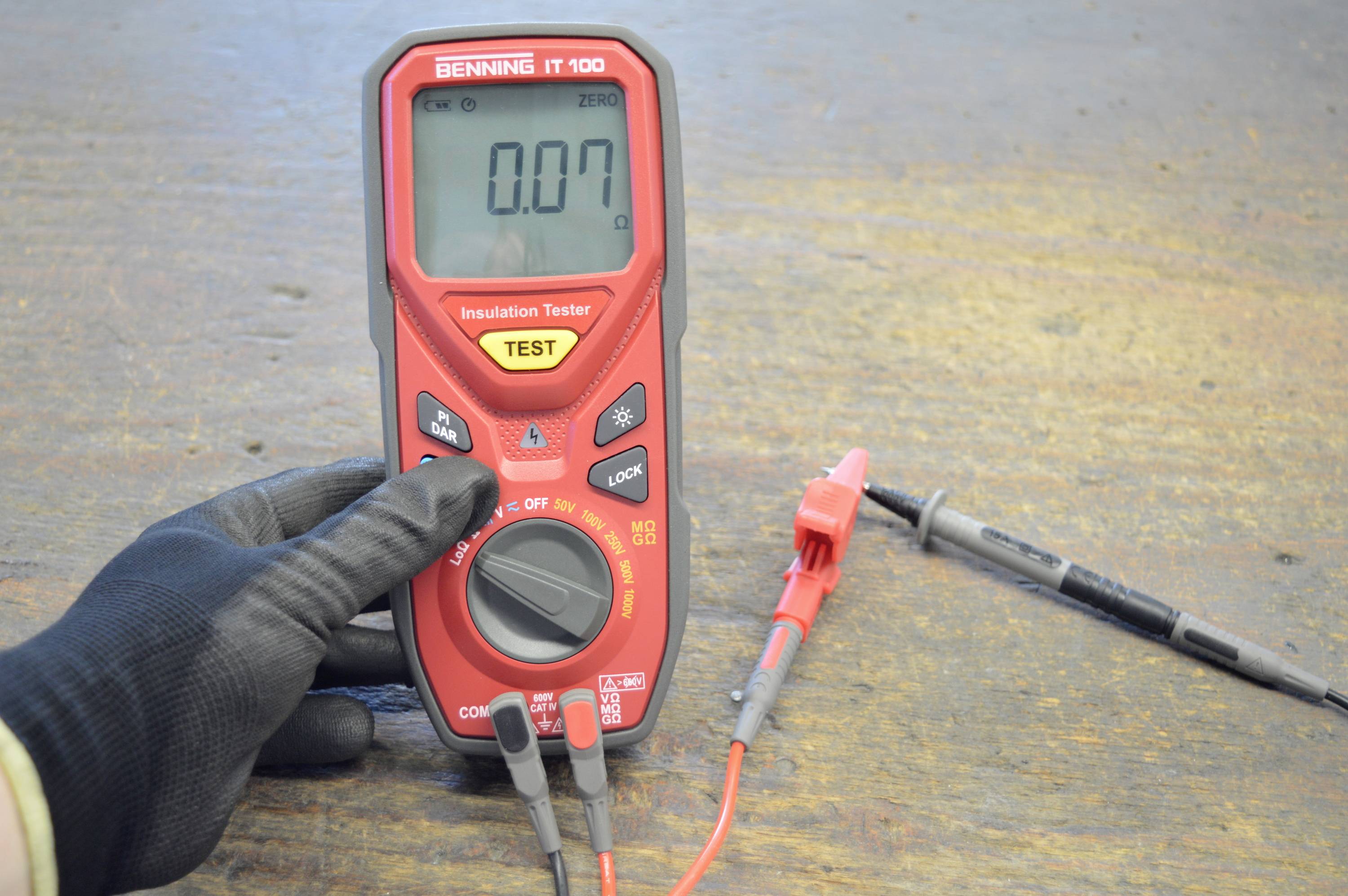 Een rood multimeter geeft '0.07' aan. Een gehandschoende hand houdt het apparaat vast, dat is aangesloten op twee kabels met klemmen.