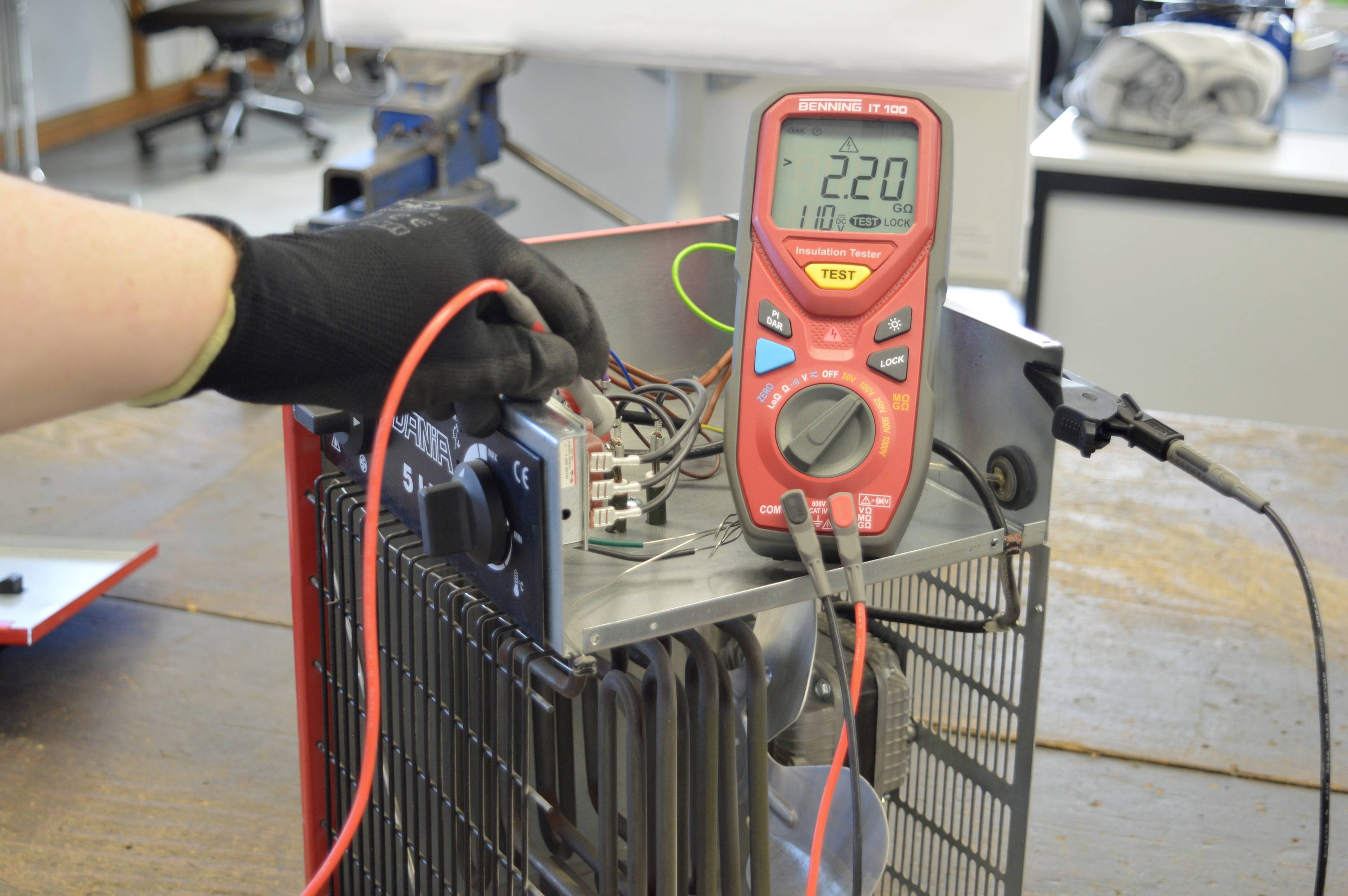 Een hand met een zwarte handschoen houdt een multimeter vast die is aangesloten op een elektrisch apparaat. Het display toont '220' volt.