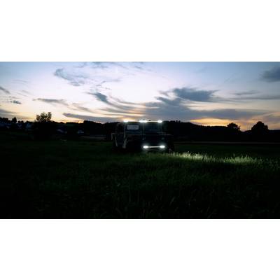 Een voertuig met brandende koplampen staat bij schemering op een weiland. De lucht vertoont een kleurrijke zonsondergang.