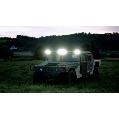Een militair voertuig met brandende koplampen staat op een weide bij schemeering, omgeven door bomen en heuvels.