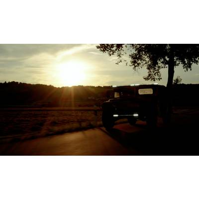 Een Jeep rijdt bij zonsondergang langs een landweg, omgeven door velden en heuvels op de achtergrond.