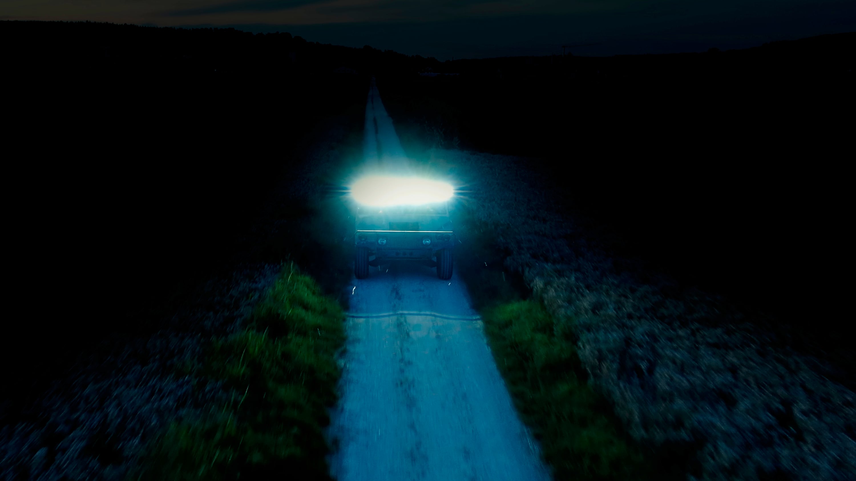 Een voertuig met fel koplamplicht rijdt 's nachts op een verlaten weg. De omgeving is donker en onverlicht.