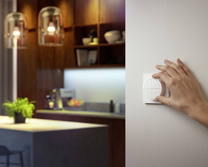 Een hand draait aan een slimme lichtdimmer aan de muur in een gezellige woonkamer met een keukenruimte op de achtergrond.