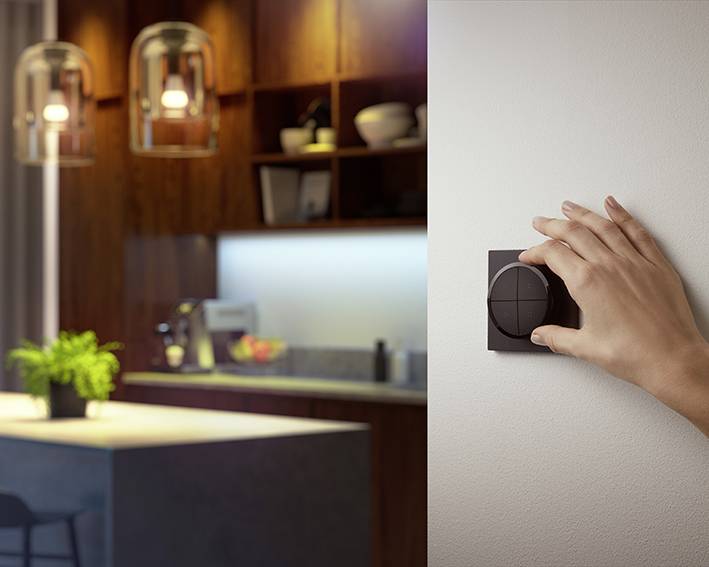 Een hand draait een lichtdimmer aan de muur. Op de achtergrond een modern verlichte keuken met planten en keukengerei.