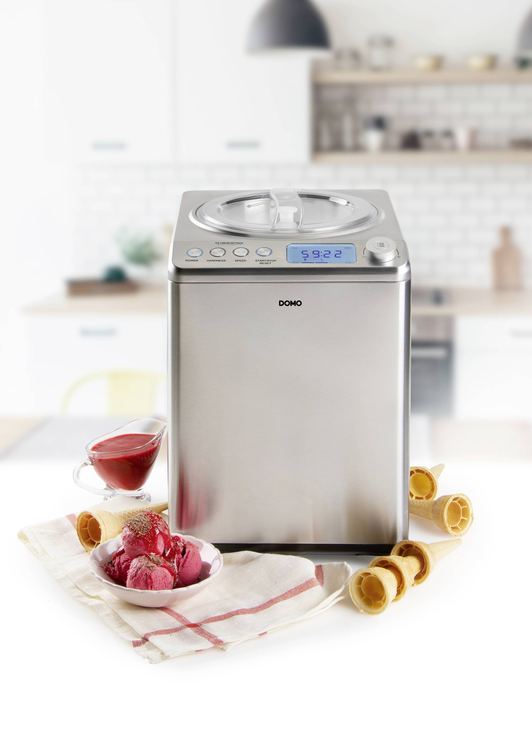 Een zilveren ijsmachine staat op een tafel in een keuken. Ernaast een kom met rood ijs en lege ijshoorntjes.