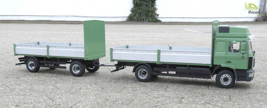 Een groene model-vrachtwagen met aanhanger staat op een geasfalteerd terrein. Op de achtergrond zijn een witte muur en planten zichtbaar.