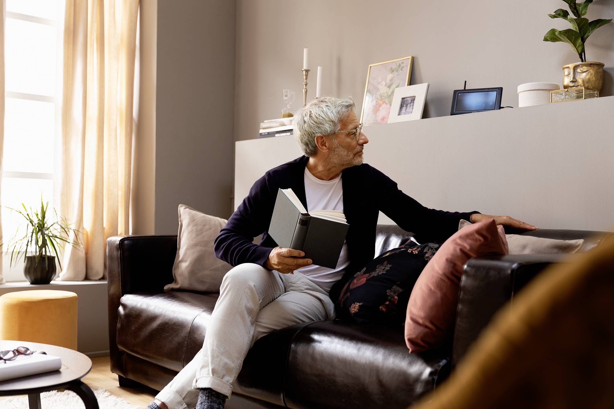 Een oudere man zit op een bank in een woonkamer, houdt een boek vast en kijkt nadenkend opzij.