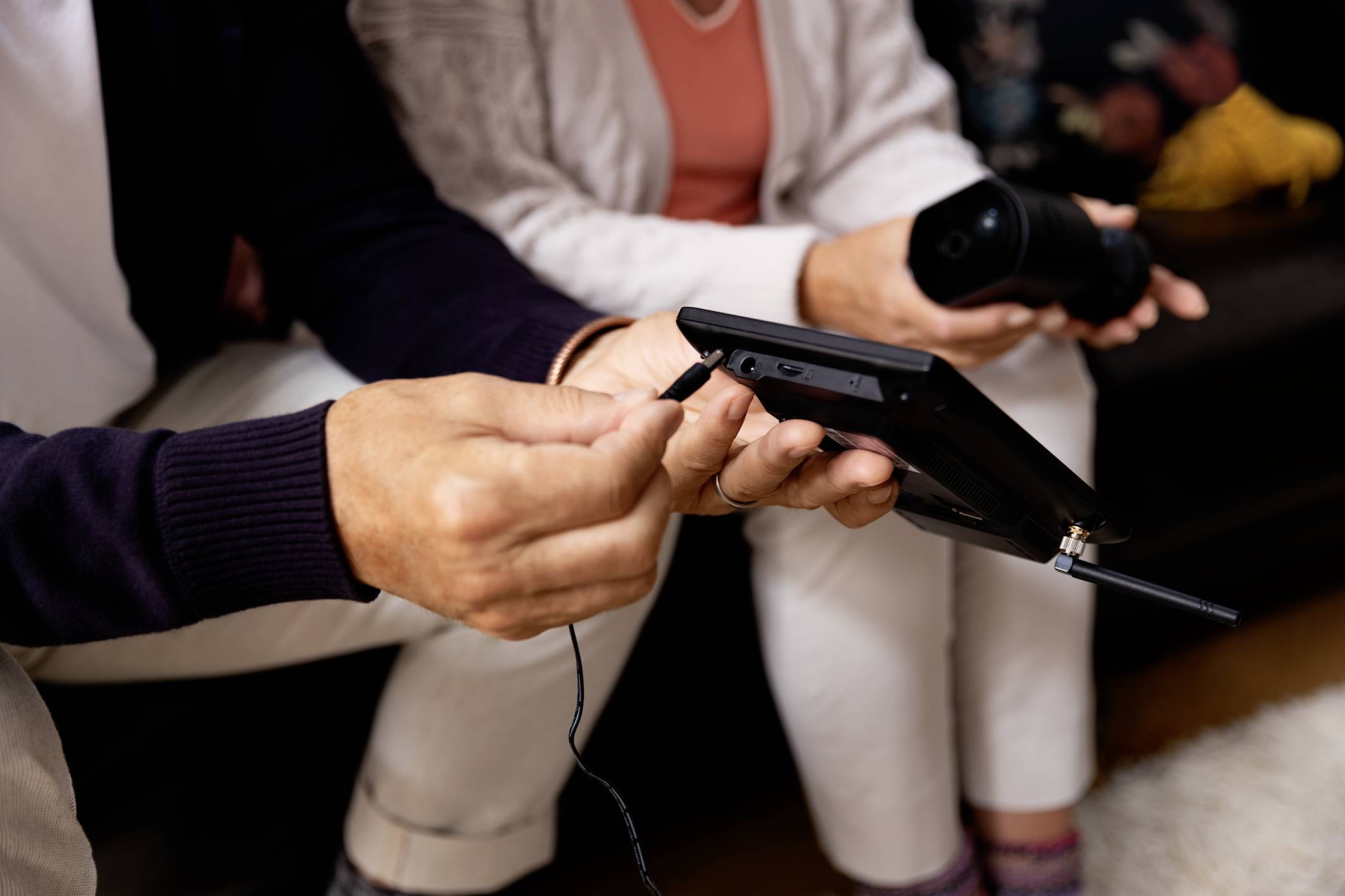 Een persoon laadt een tablet op, terwijl een andere persoon ernaast zit en een beker vasthoudt.