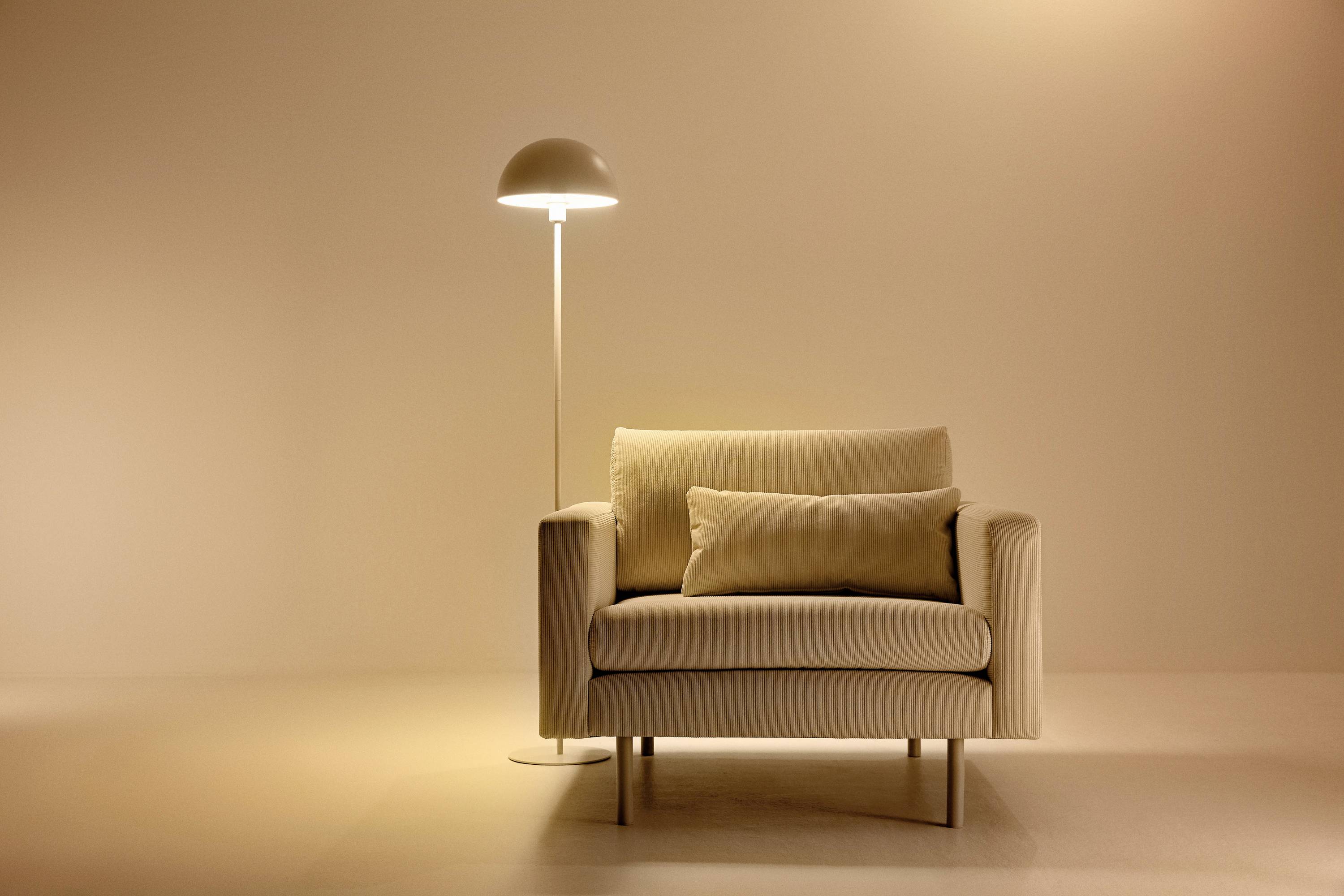 Een beige fauteuil met een kussen staat naast een moderne vloerlamp in een minimalistisch ingerichte ruimte.