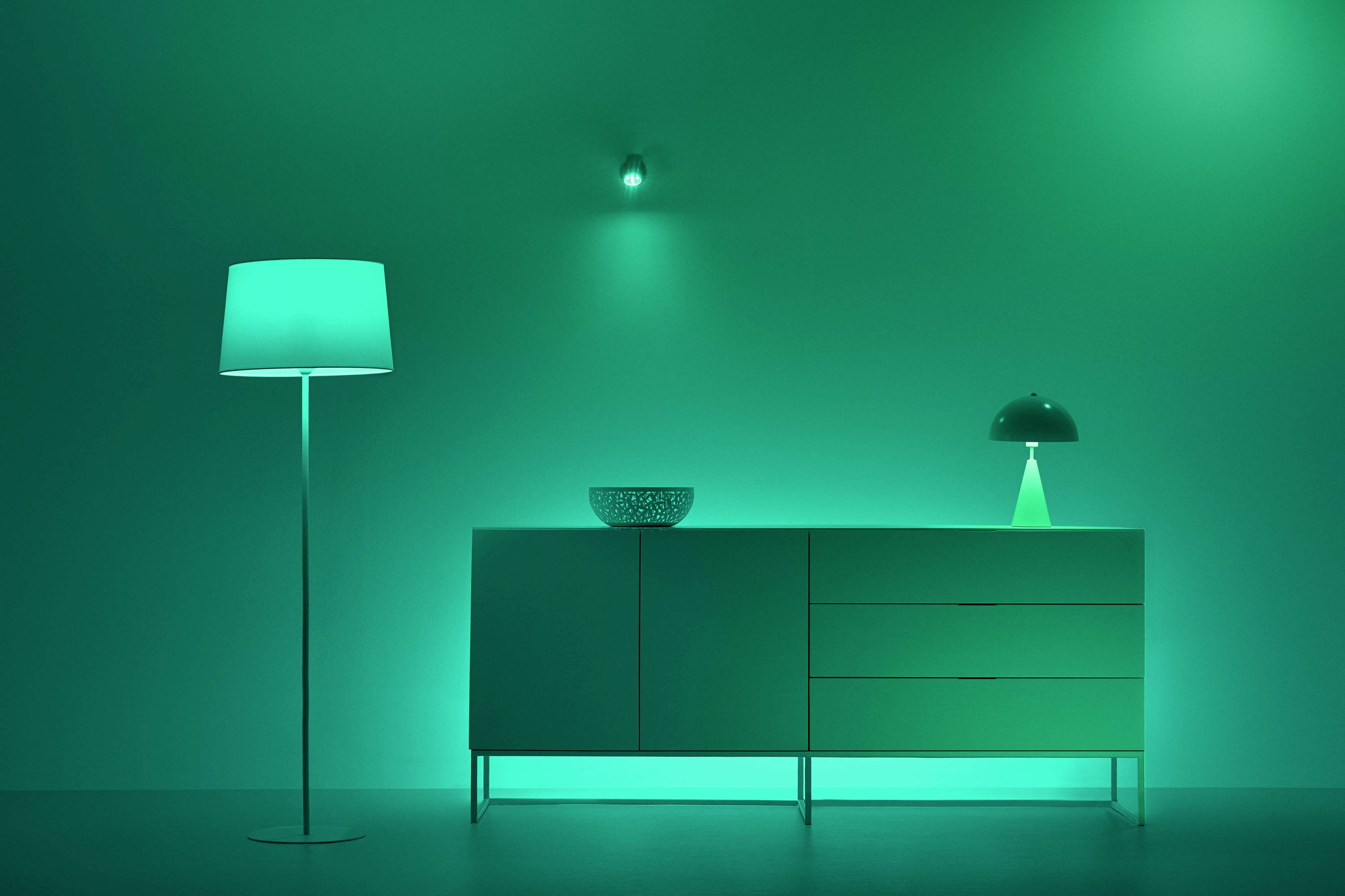 Een ruimte met groen licht, waarvan links een staande lamp staat, in het midden een kast met een kom erop en rechts een tafellamp.