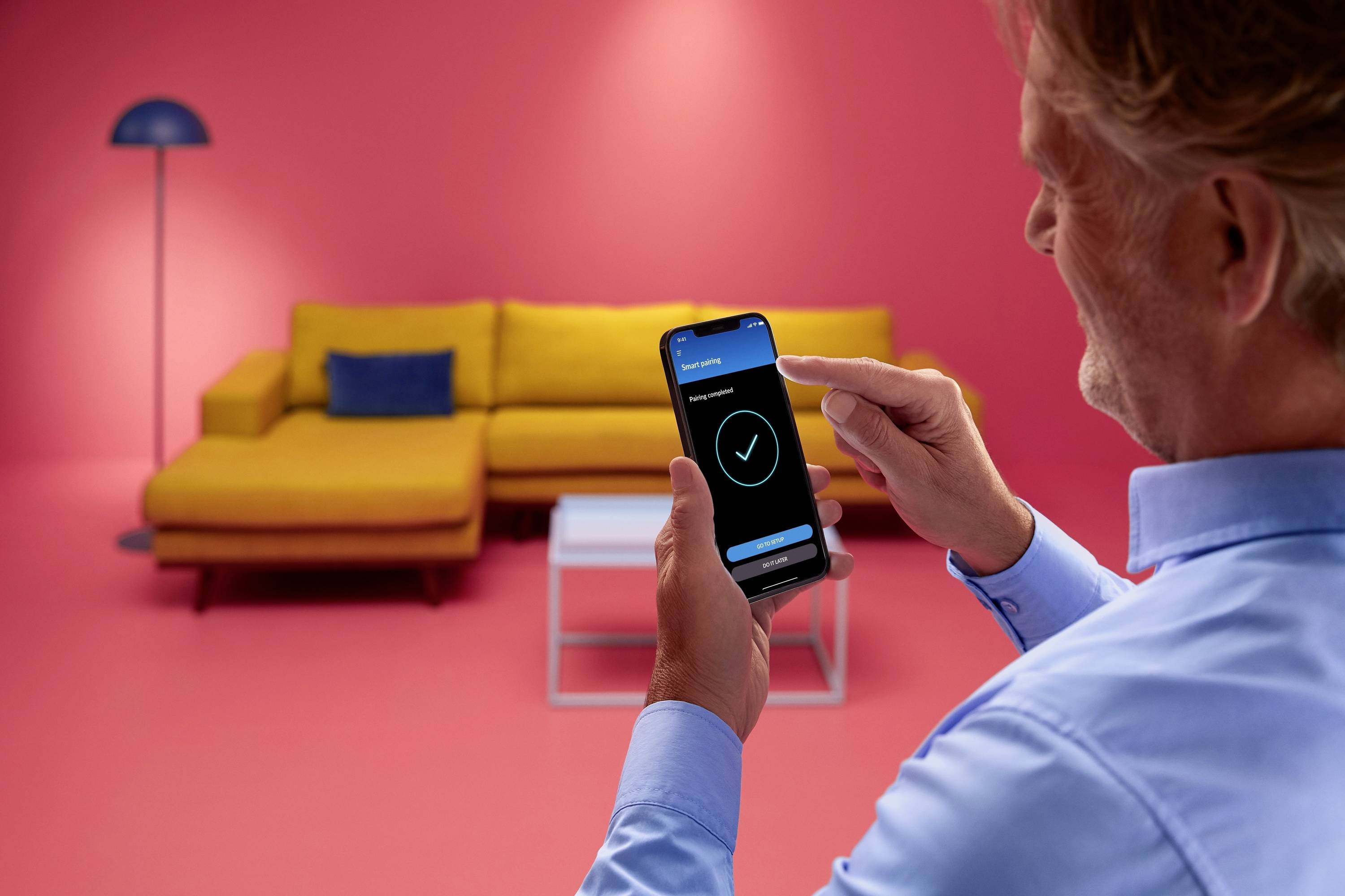 Een persoon bedient een smartphone met een app in een ruimte met een gele bank en roze muren. Een witte tafel en een lamp zijn zichtbaar.