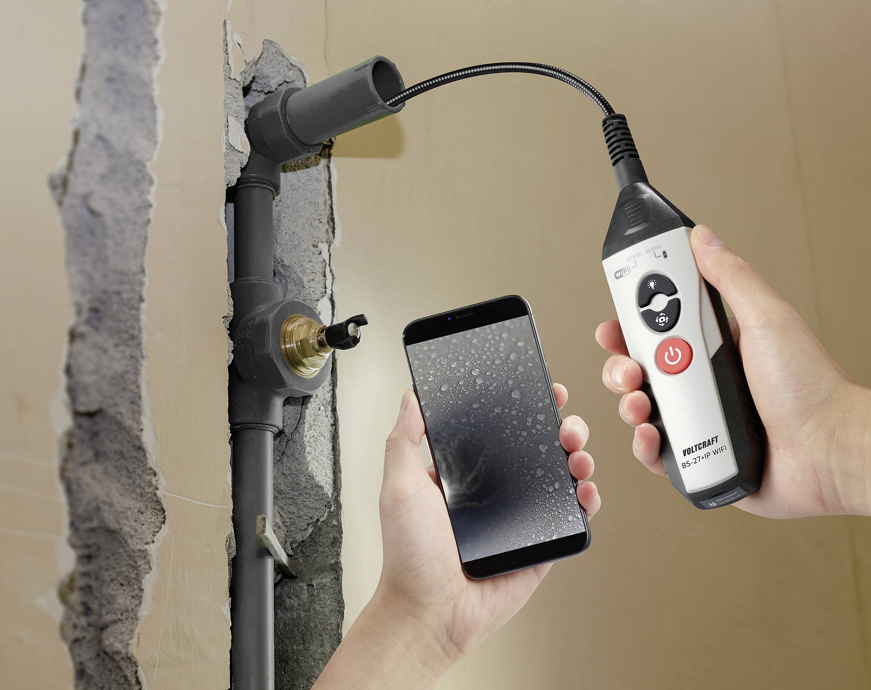 Een hand houdt een endoscoopinstrument vast dat een waterleiding inspecteert door een opening in de wand. De andere hand houdt een smartphone vast waarop het endoscopische beeld wordt weergegeven.