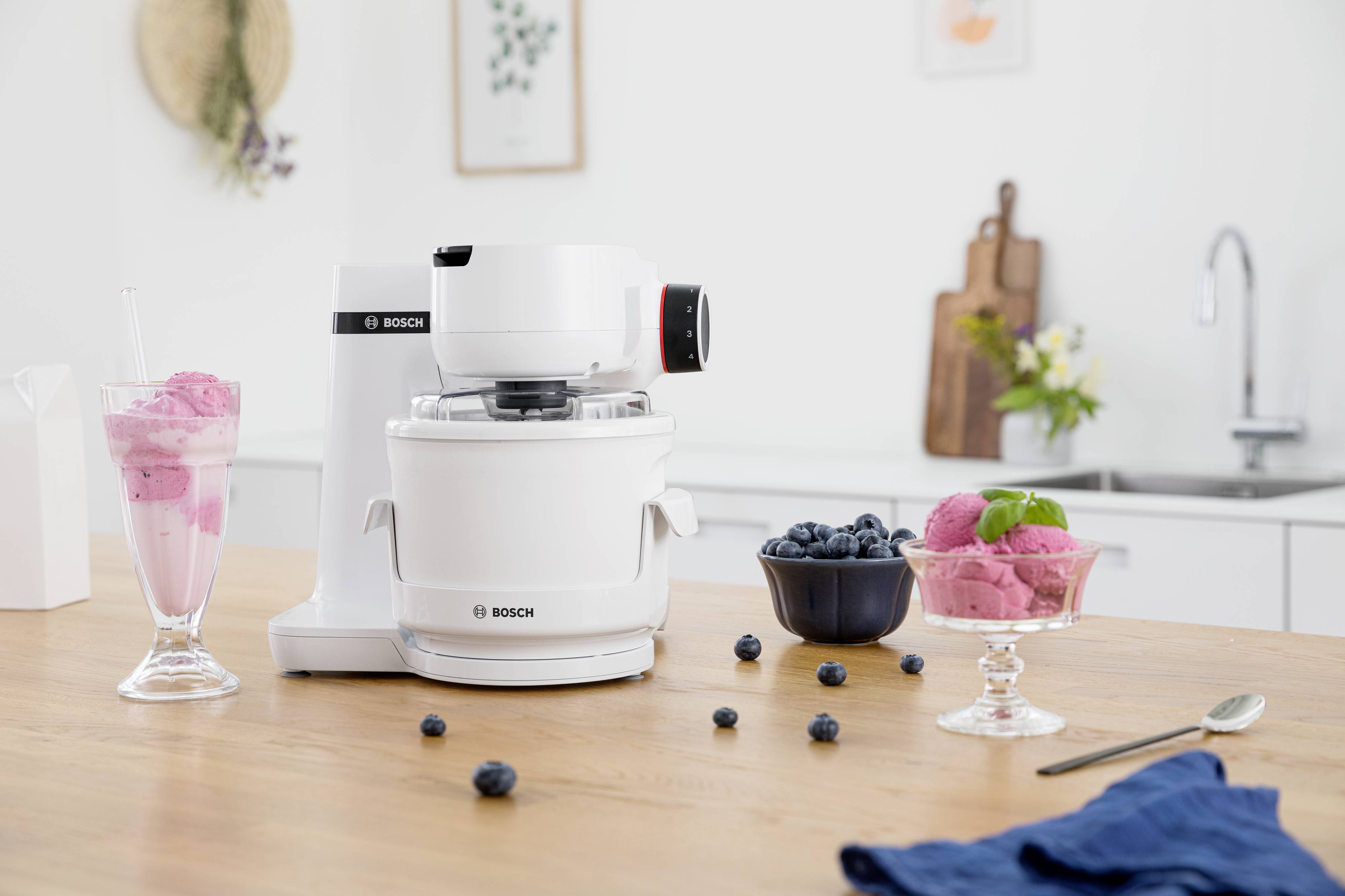 Een keukenmachine op een tafel, omringd door een milkshake, een kom met bosbessen en een dessert met roze ijs.