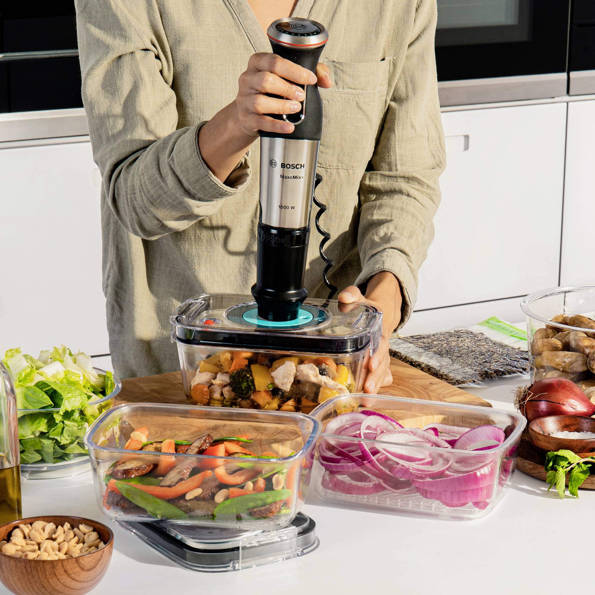 Een persoon gebruikt een staafmixer om groente in een kom te pureren. Op tafel liggen verschillende voedingsmiddelen en ingrediënten.
