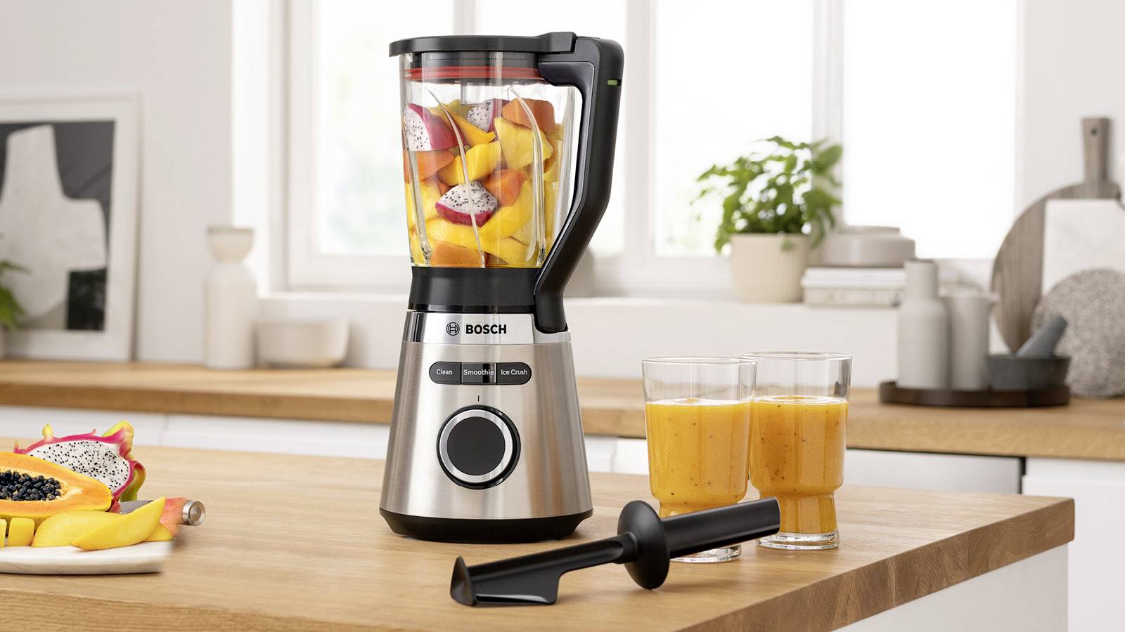 Een zilveren staande blender op een aanrecht, gevuld met kleurrijk fruit. Ernaast staan twee glazen met oranje smoothie.
