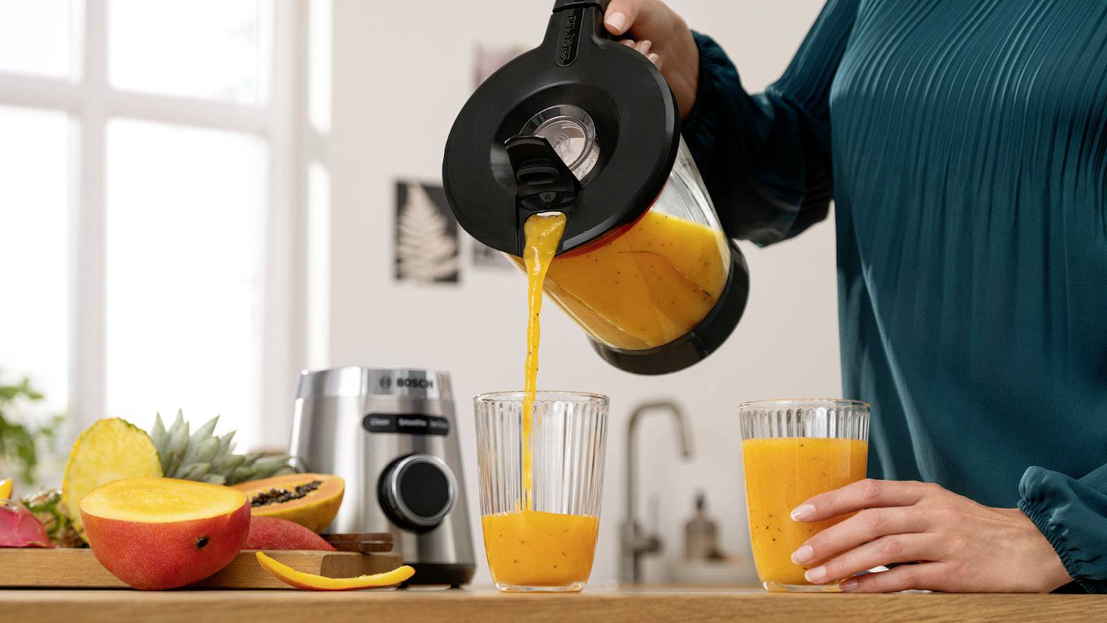 Een persoon giet verse sinaasappelsap uit een blender in twee glazen. Op de achtergrond zijn verschillende soorten fruit te zien.