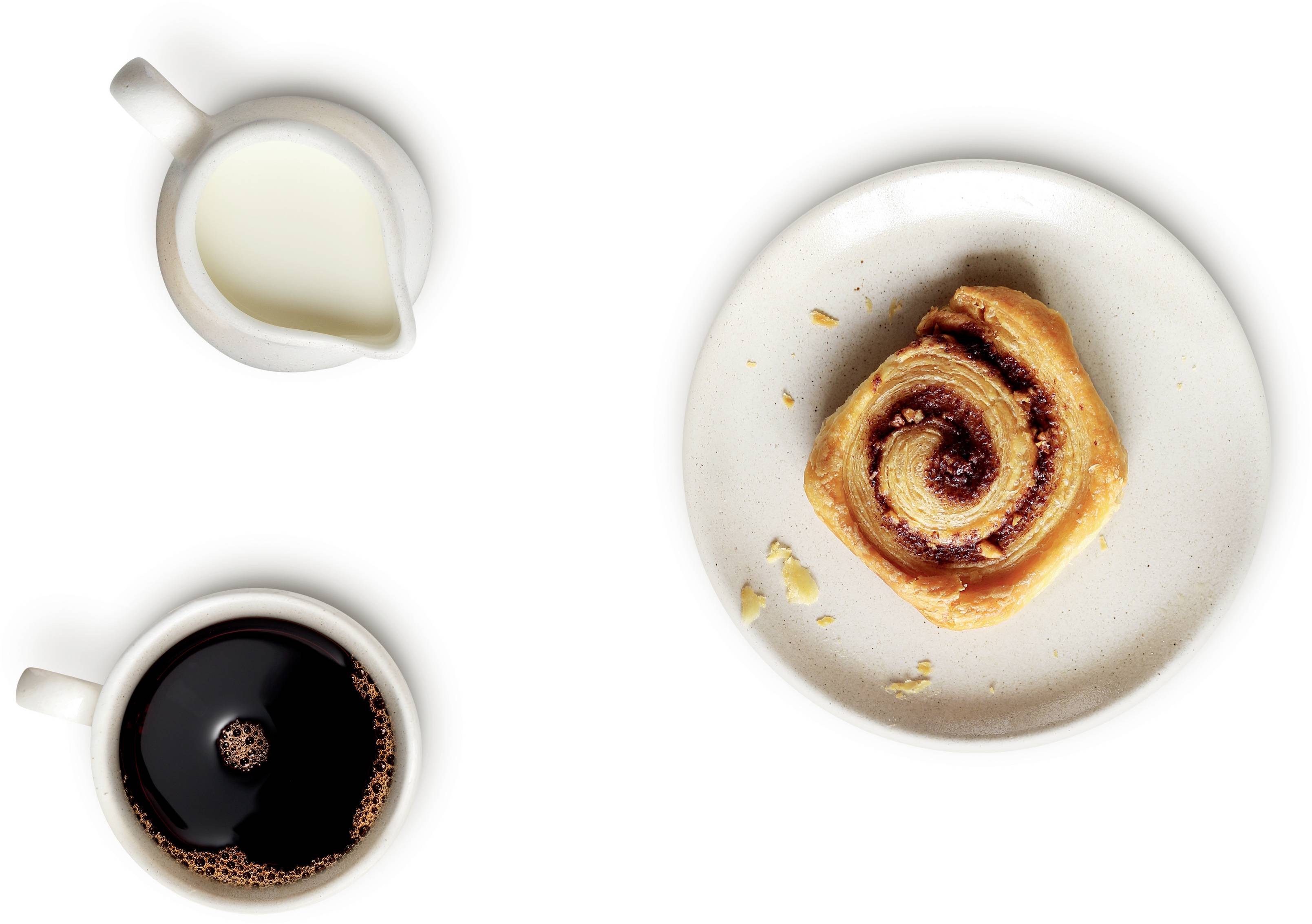 Kopje koffie, melkkannetje en een kaneelbroodje op een bord.