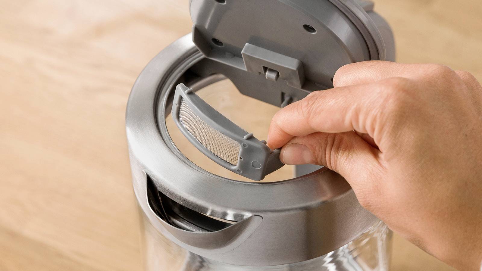 Een hand opent het deksel van een zilveren waterkoker met ingebouwd filter, op een houten oppervlak.