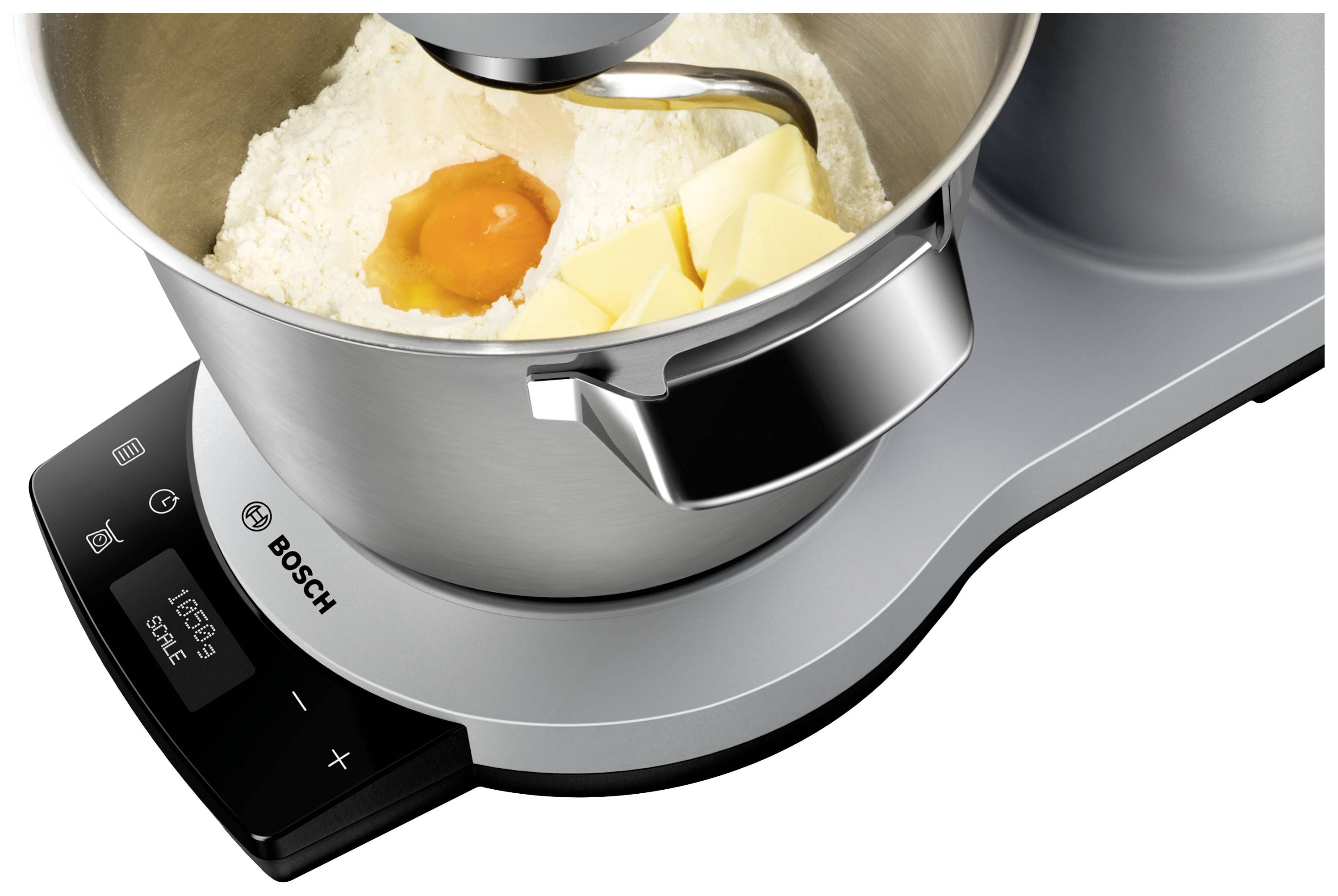 Een keukenmachine kneedt ingrediënten in een kom. Zichtbaar zijn bloem, eigeel en blokjes boter. De machine is in bedrijf.