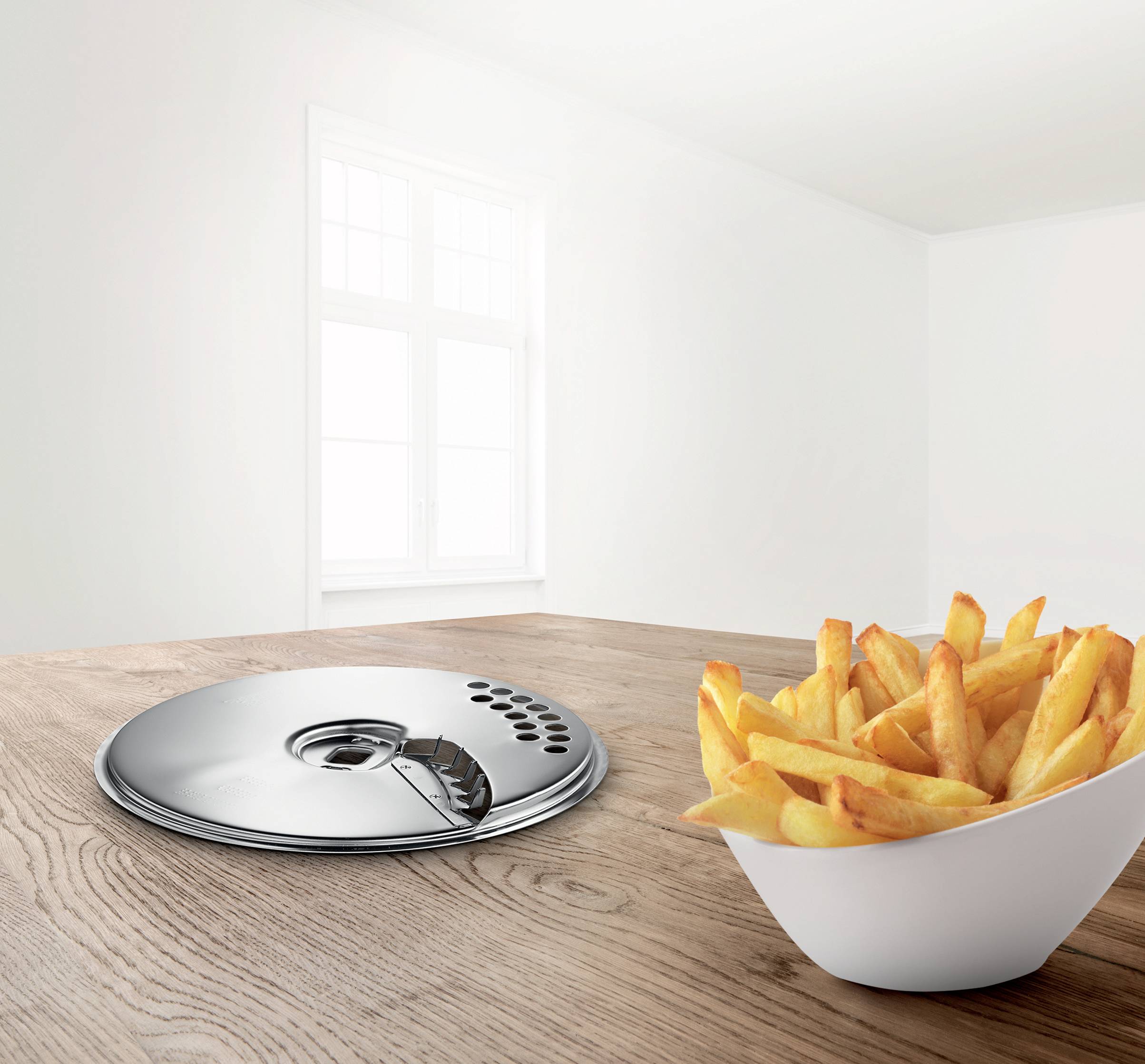 Een witte kom met frietjes staat op een houten tafel naast een plat, rond zilverkleurig voorwerp, met op de achtergrond een raam.