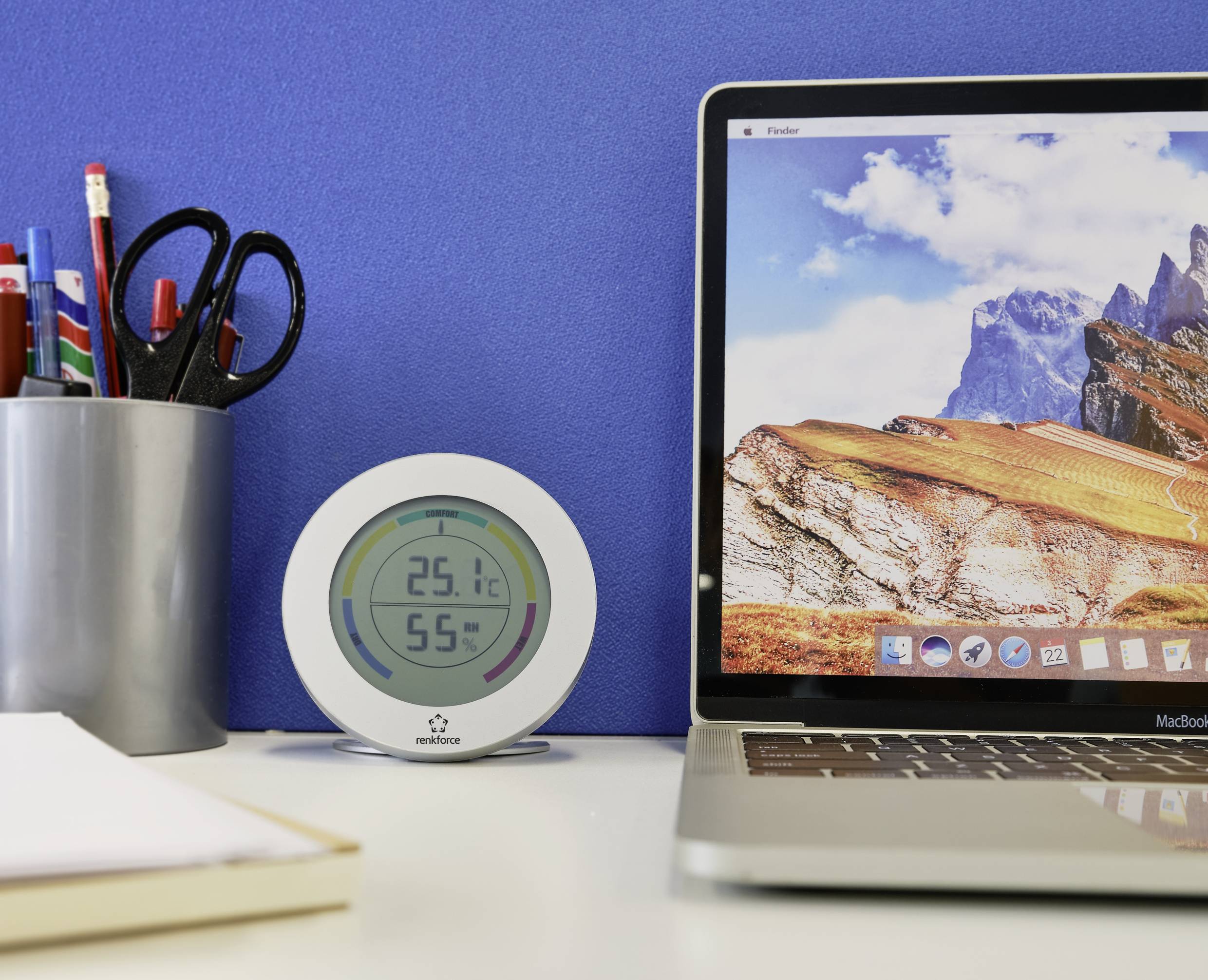 Op een bureau staan een laptop met een bergachtergrond, een ronde digitale thermometer die 25,1°C en 55% luchtvochtigheid aangeeft, en een beker met pennen.