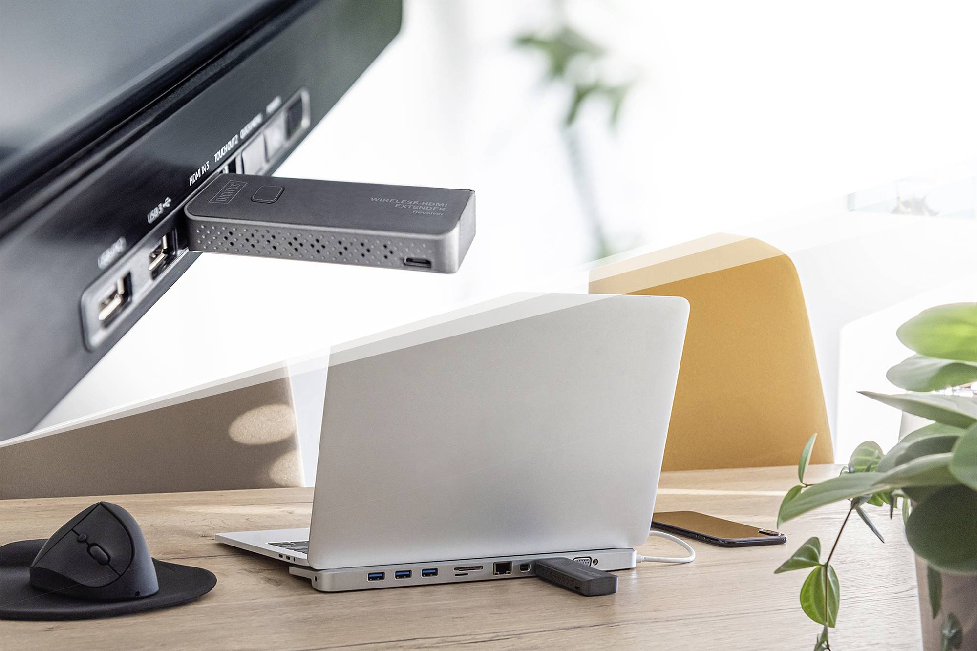 Een laptop staat op een bureau met een dockingstation en toebehoren. Een televisie vertoont een aangesloten streamingbox. Planten op de achtergrond.