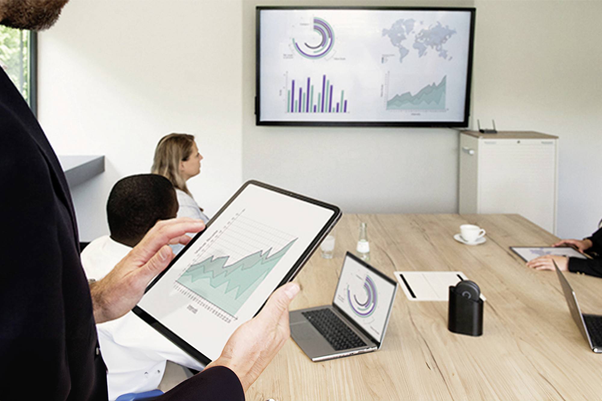 Personen in een vergaderzaal bestuderen diagrammen op tablets en laptops, terwijl een groot scherm grafische informatie weergeeft.