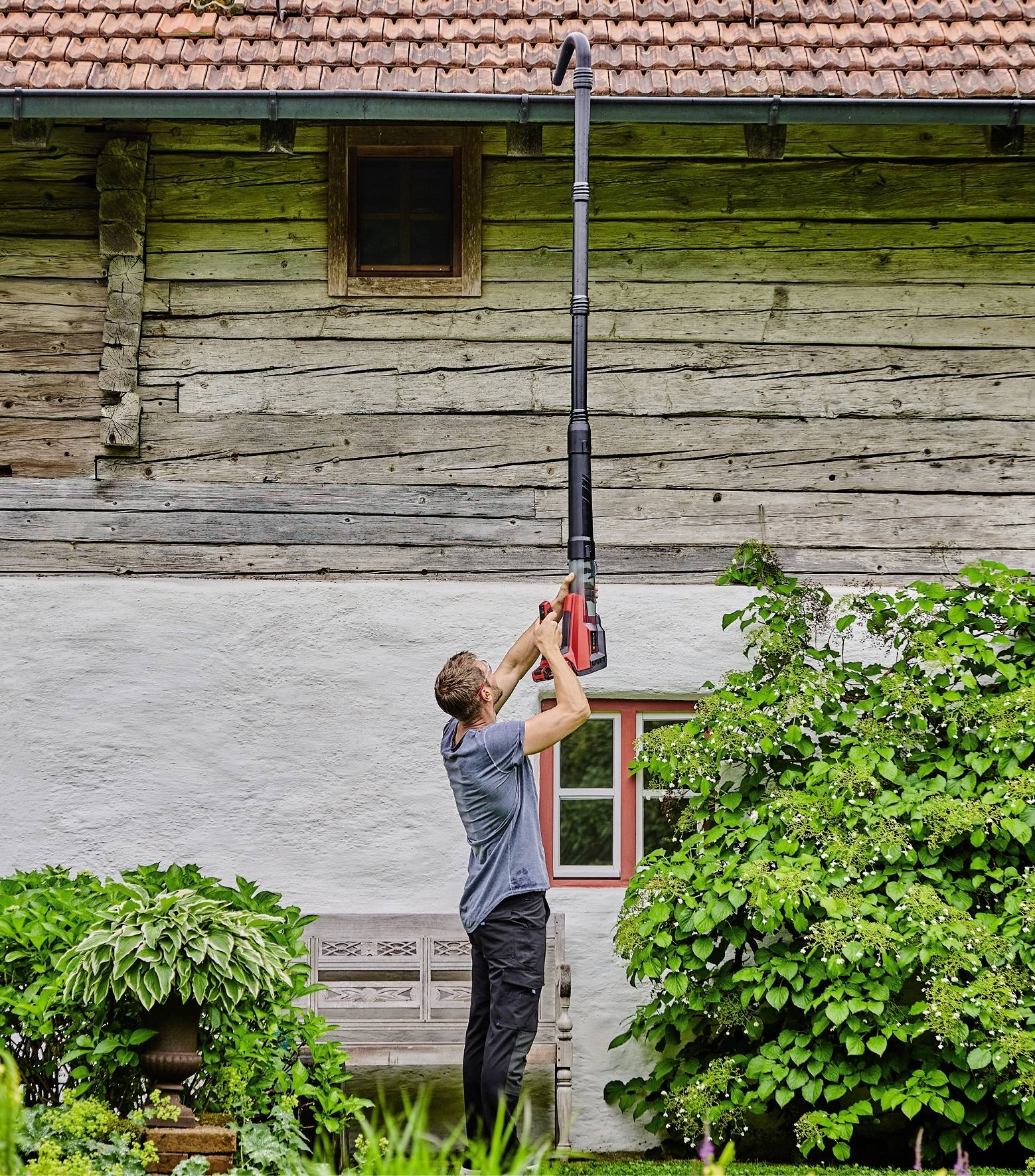 Een man gebruikt een lange bladblazer om het dak van een oud houten huis schoon te maken. Op de voorgrond bevinden zich groene struiken en planten.