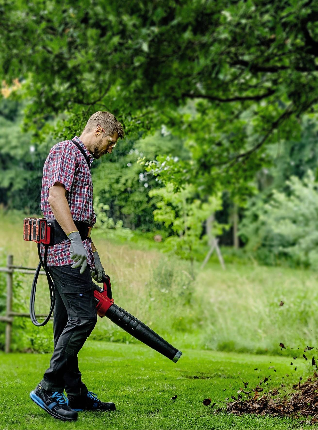 Een man in de tuin blaast bladeren met een bladblazer. Hij draagt tuinhandschoenen en ziet er geconcentreerd uit. De achtergrond is groen en bebost.