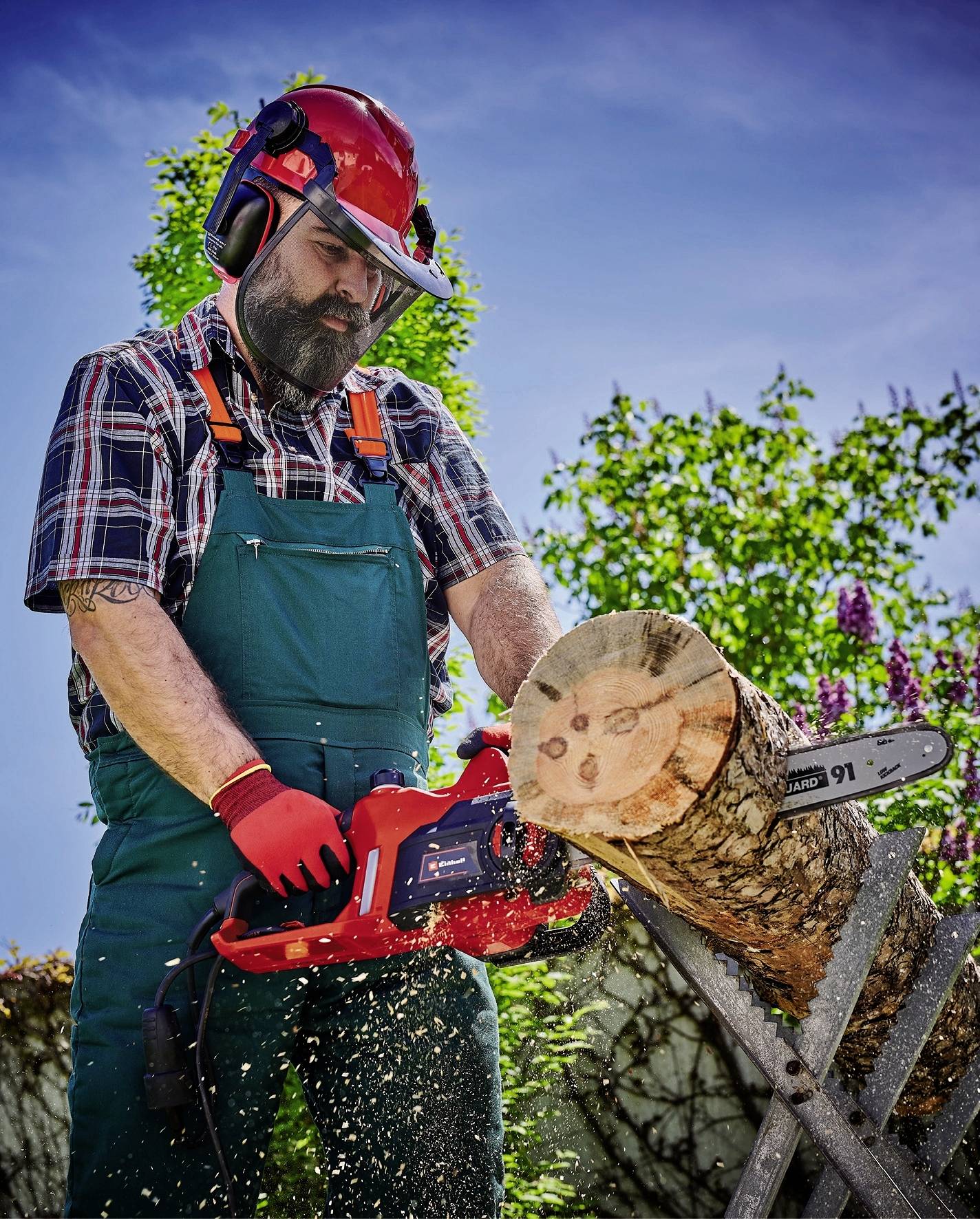Man met beschermende kleding zaagt met een kettingzaag een boomstam op een zaagbok in de buitenlucht onder een heldere hemel.