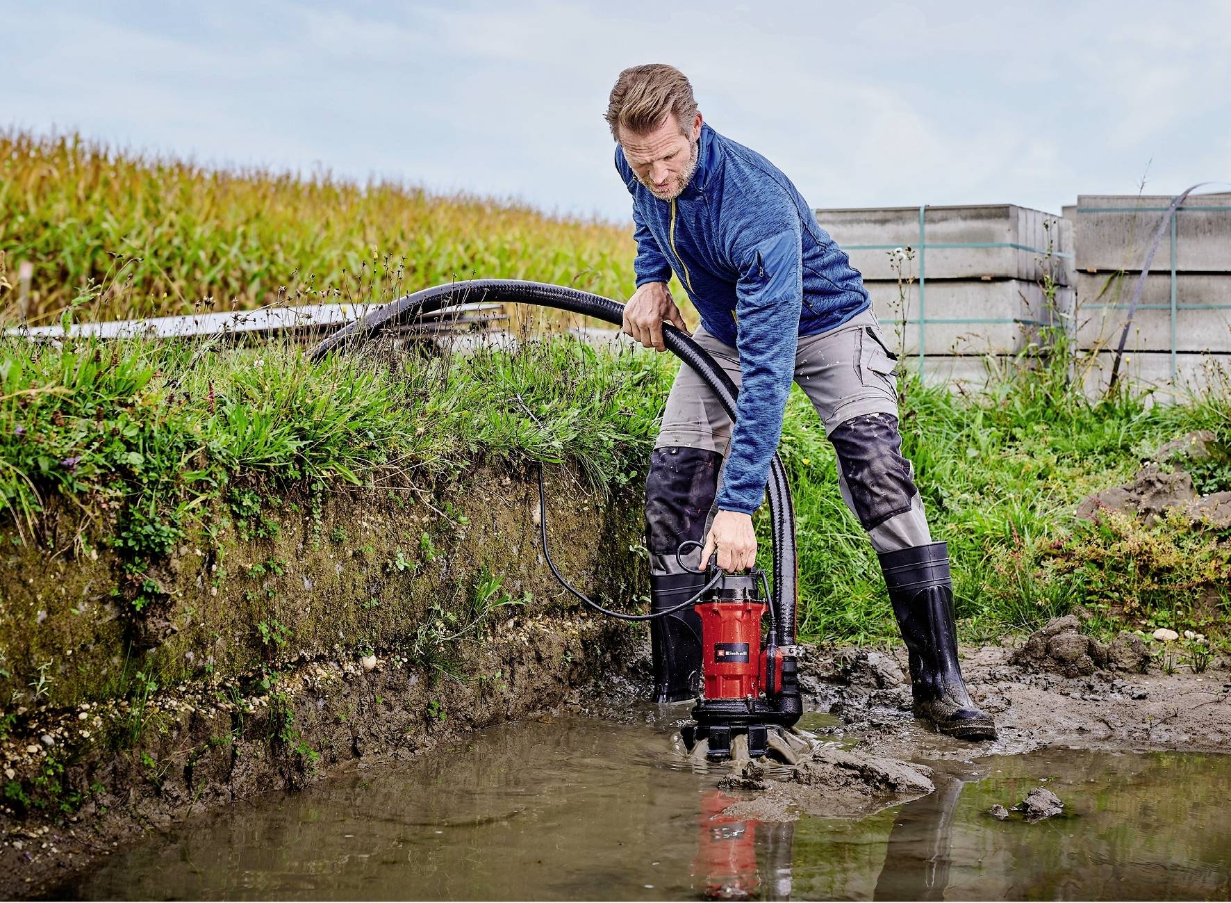 Een man in werkkleding pompt water uit een sloot met een kleine, rode waterpomp. Op de achtergrond is een veld zichtbaar.