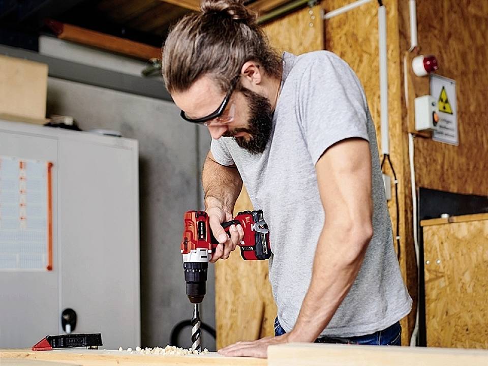 Een man met veiligheidsbril werkt met een accuschroevendraaier aan een houten plank in een werkplaatsomgeving.