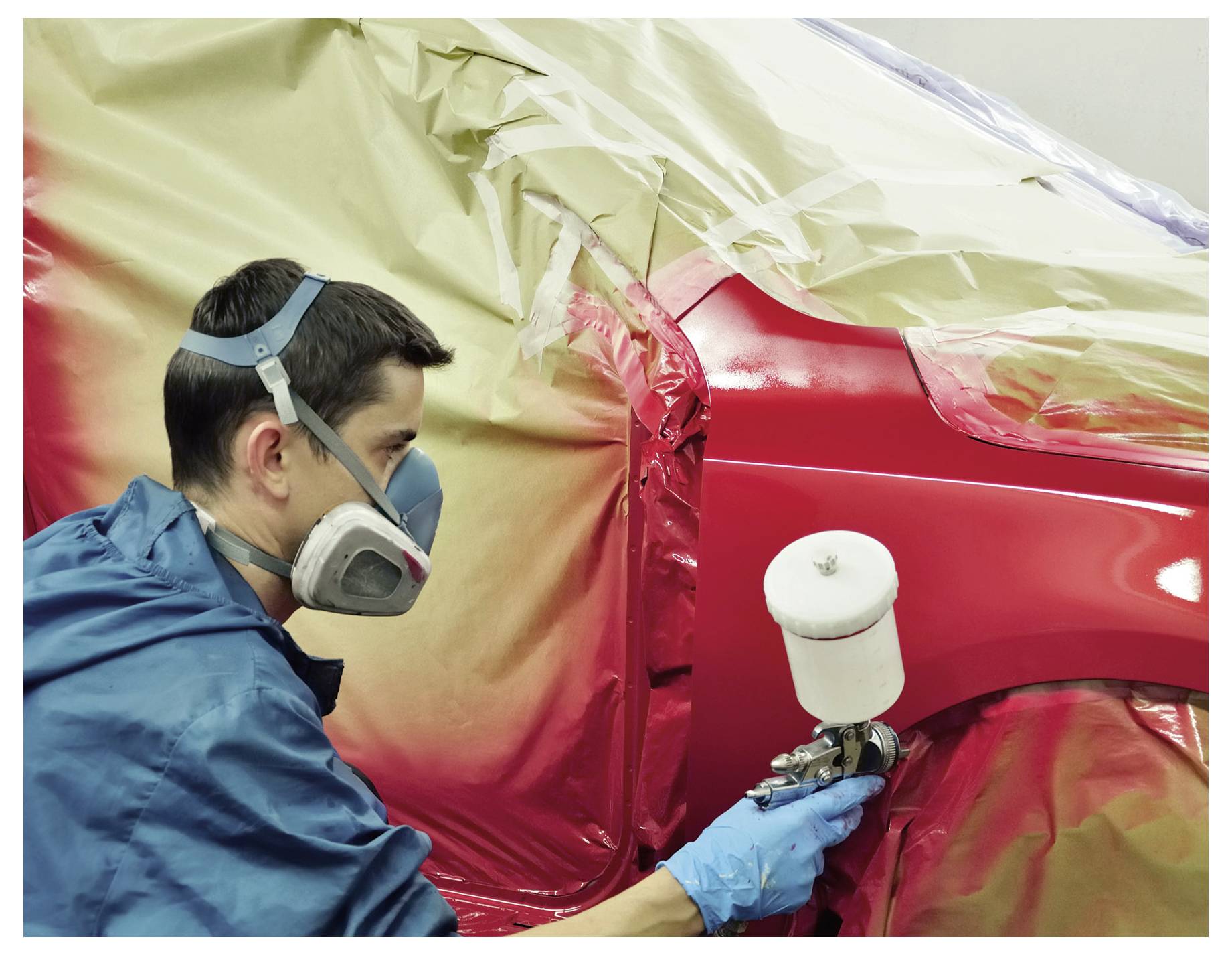 Een technicus in masker en handschoenen spray-verft een rood autodeel, met aangrenzende gebieden afgedekt met plastic folie.