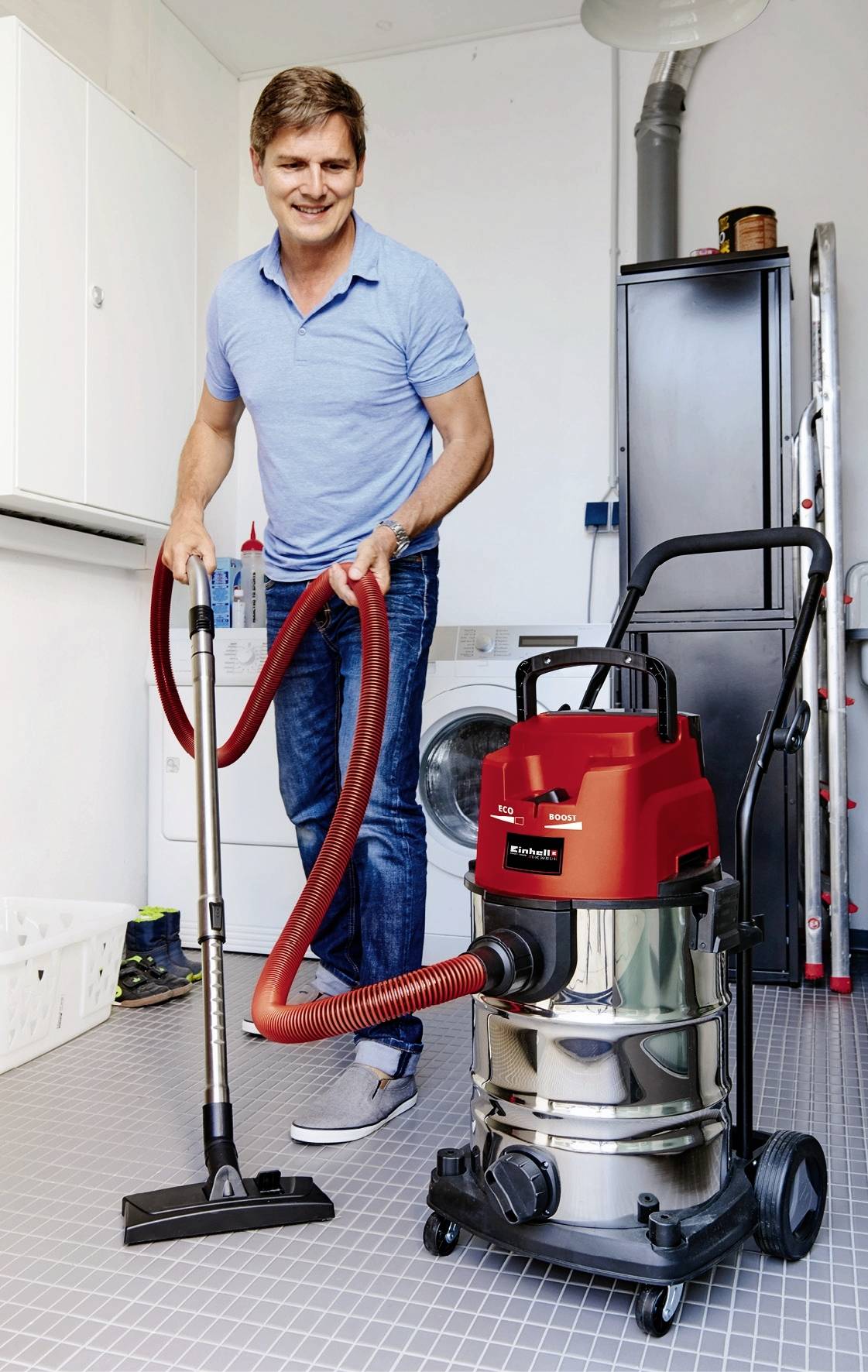 Een man glimlacht terwijl hij een industriële stofzuiger gebruikt in een bijkeuken. Op de achtergrond zijn huishoudelijke apparaten zichtbaar.