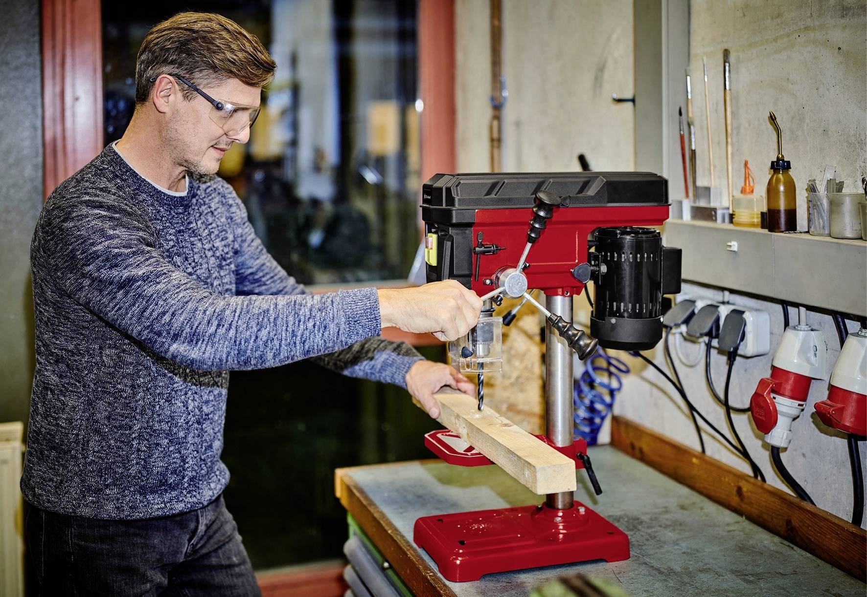 Een man werkt aan een rode kolomboormachine in een werkplaats. Hij boort een gat in een stuk hout, omringd door gereedschappen.
