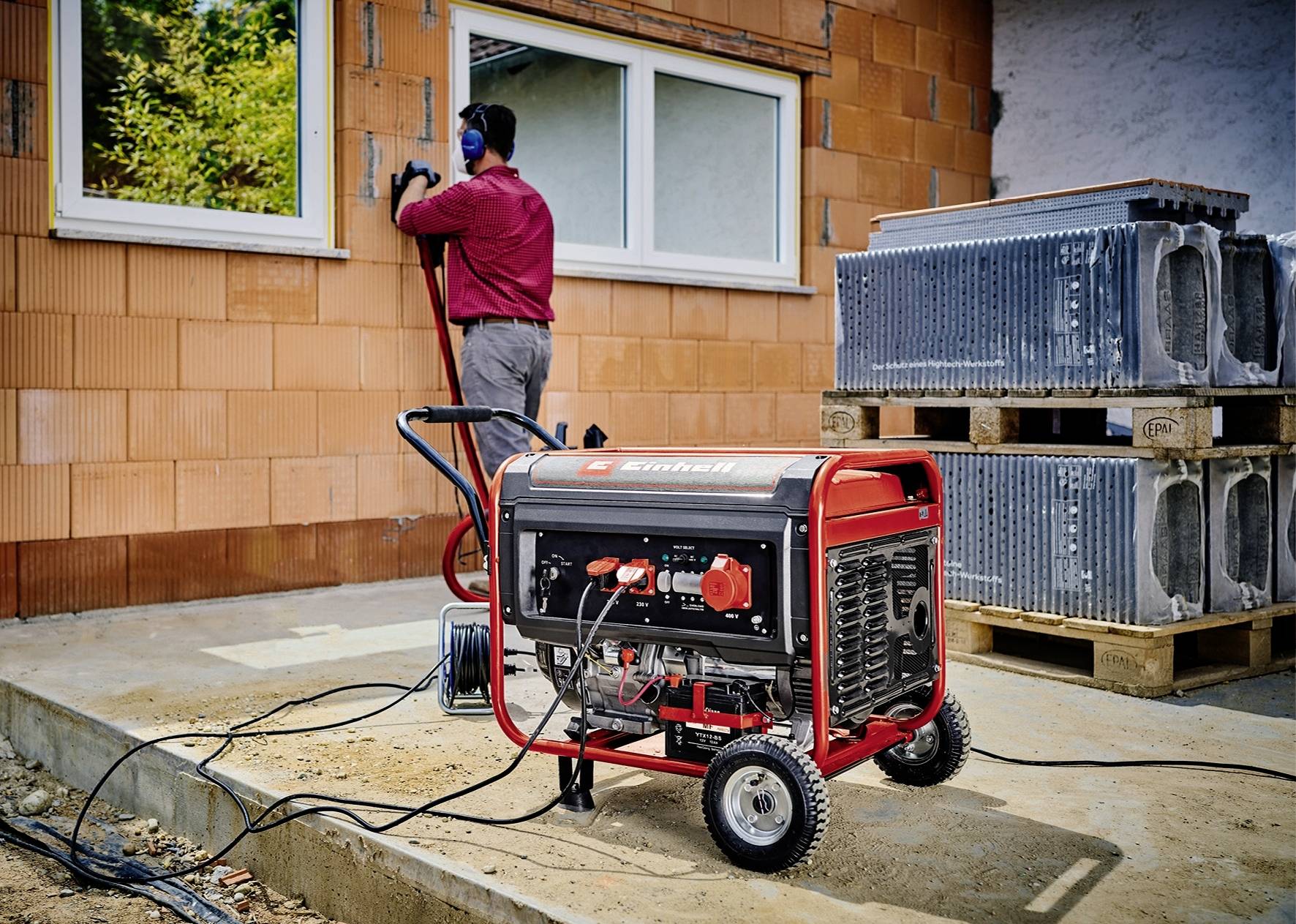 Een draagbare generator staat vooraan op een bouwplaats. Op de achtergrond werkt een persoon aan een onbepleisterde muur van een huis.