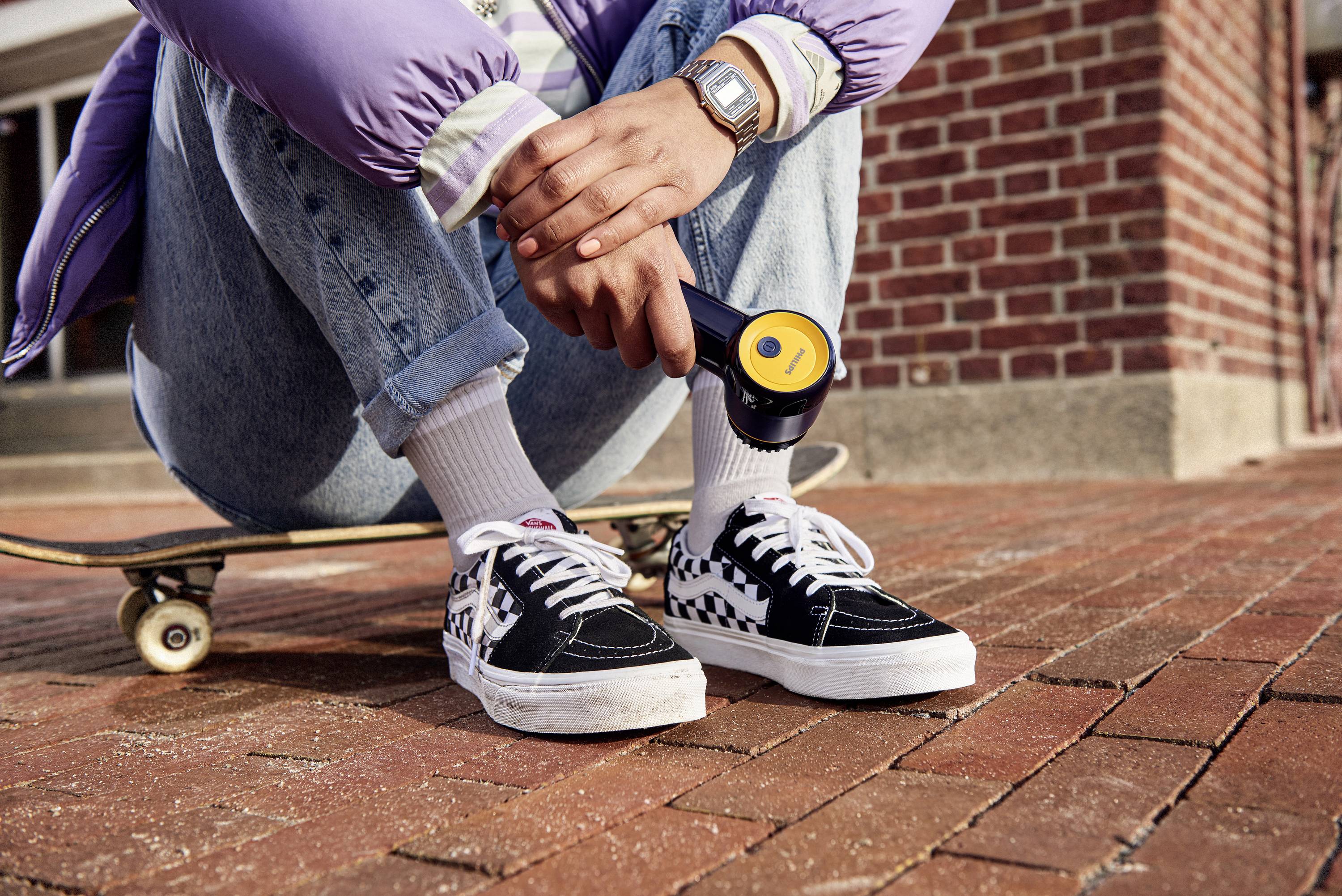 Een persoon zit en houdt een zwart voorwerp voor een skateboard op een straatweg met klinkers. Focus op schoenen en handen.