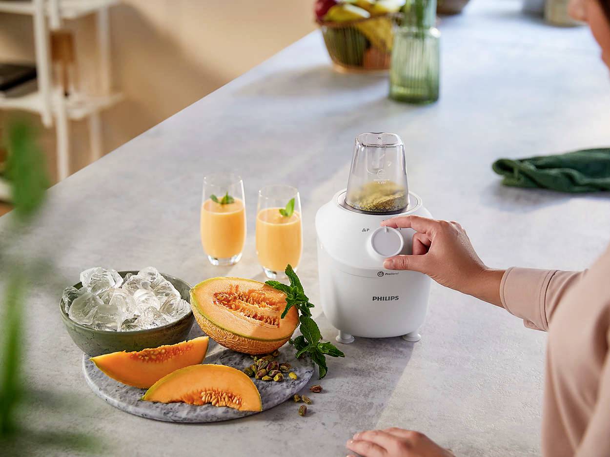 Een persoon bedient een keukenmixer op het aanrecht. Ernaast liggen meloenplakken en twee glazen met een oranje drankje.