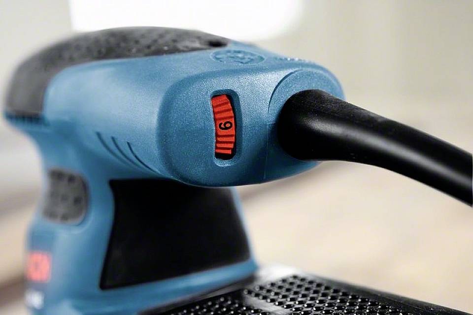 Een blauwe elektrische slijpmachine met een zwarte handgreep en een rode snelheidsregelaar in stand 6, ligt op een houten tafel.