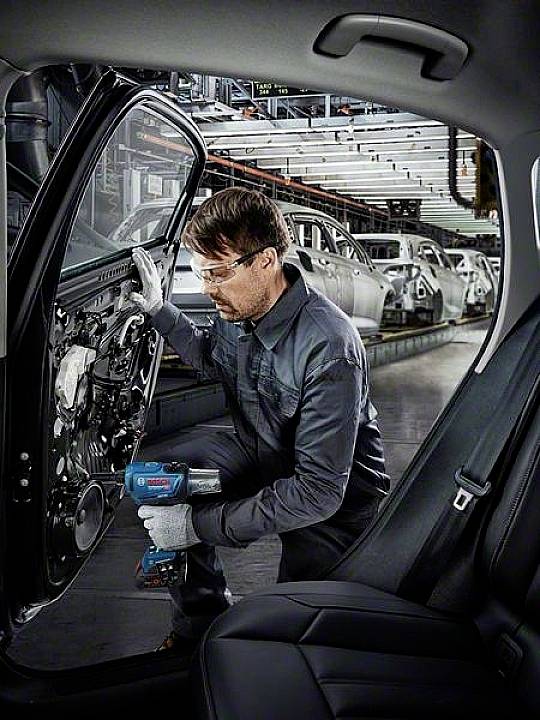 Een arbeider in een fabriek monteert een autoportier met een elektrische schroevendraaier. Op de achtergrond zijn productelijnen met andere voertuigonderdelen zichtbaar.