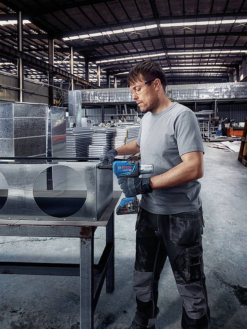 'Man in werkplaats bewerkt metalen onderdeel met elektrisch gereedschap, omringd door plaatwerkdelen en machines, draagt veiligheidsbril en handschoenen.'