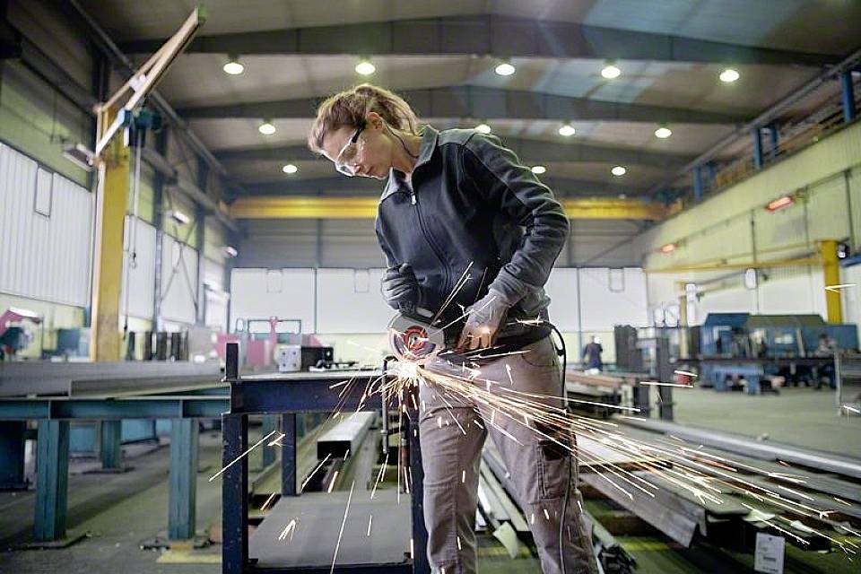 Een persoon draagt een veiligheidsbril en handschoenen en slijpt metaal met een haakse slijper in een industriële werkplaats. Vonken spatten rond.