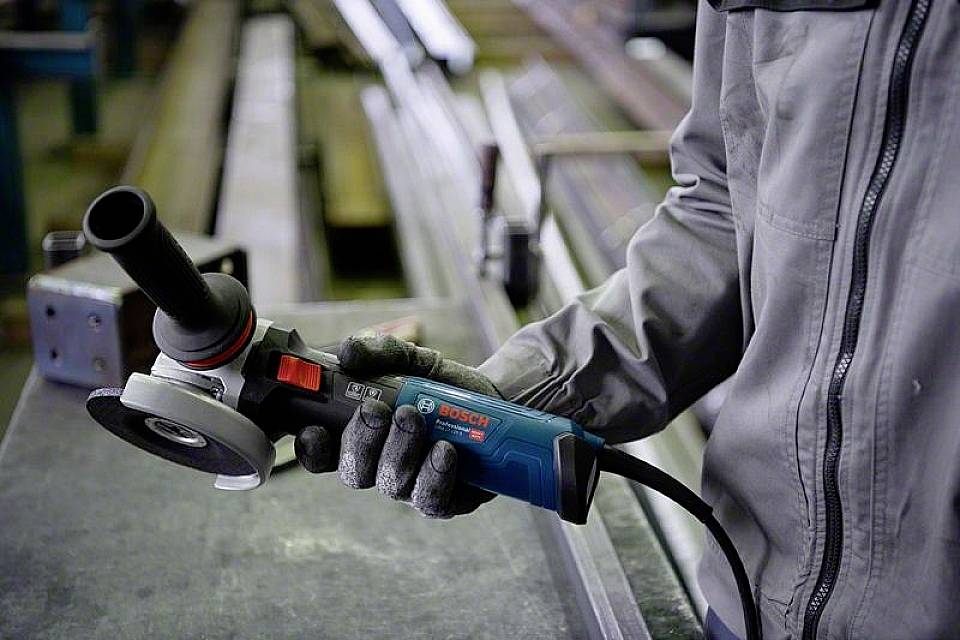 Een persoon houdt een blauwe haakse slijper vast, klaar voor gebruik in een werkplaats met metalen balken op de achtergrond.