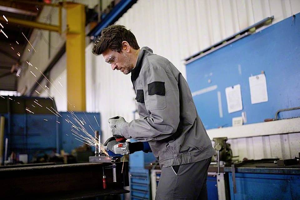 Een mens in grijze werkkleding slijpt metaal in een werkplaats. Vonken spatten rond, gereedschappen en machines bevinden zich op de achtergrond.