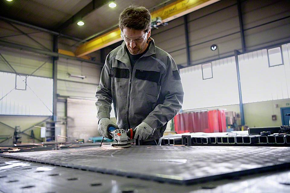 Een persoon in beschermende uitrusting slijpt metaal in een fabriekshaal. Vonken spatten van de slijpschijf, en de hal is goed verlicht.